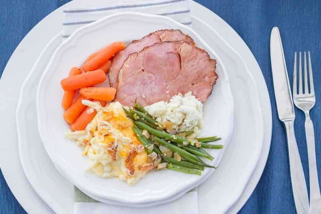 Pineapple Instant Pot Ham with Funeral Potatoes, Green Beans, and Carrots on a white plate with silver wear