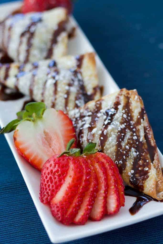 Cookie Dough Dessert Wontons with strawberries, chocolate sauce, and powdered sugar on a blue blackground