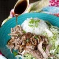 teriyaki sauce being poured onto a plate of kalua pork, rice, and cabbage on a blue plate