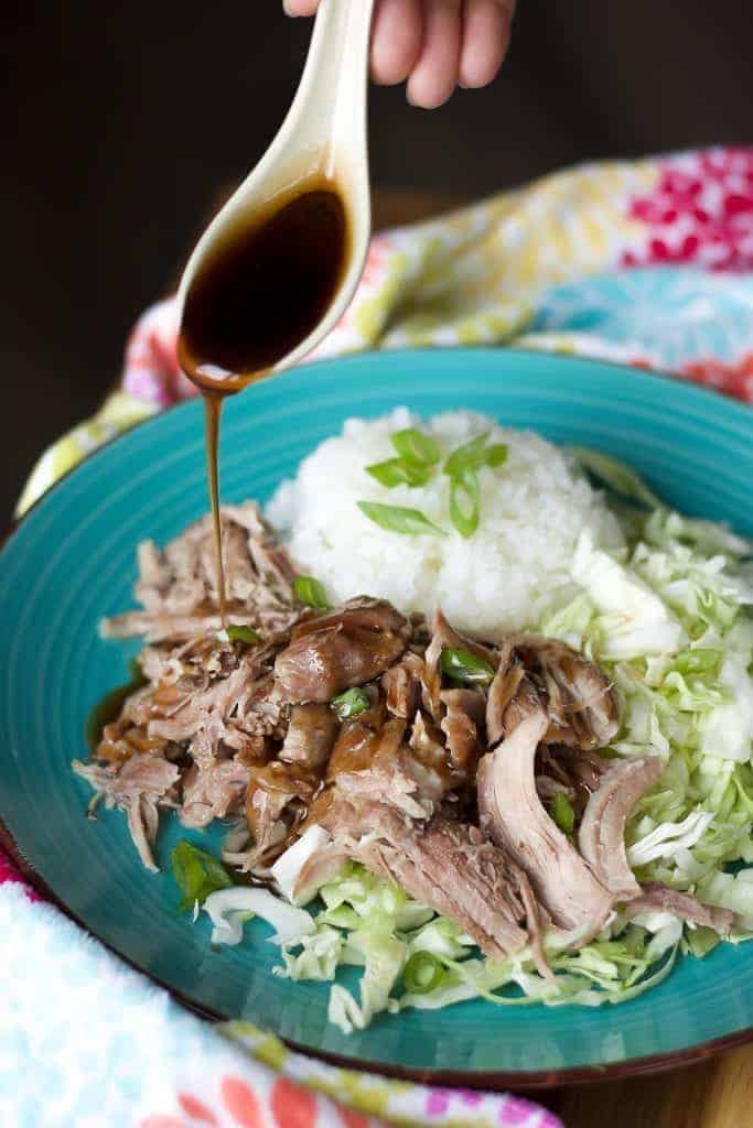 teriyaki sauce being poured onto a plate of kalua pork, rice, and cabbage on a blue plate