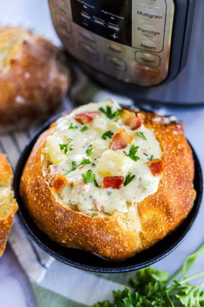  A bread bowl in a blue bowl, with clam chowder topped with bacon and parsley. Instant Pot and parsley and fresh bread in the background