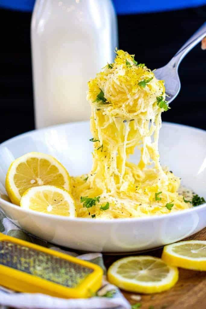 a bowl of Instant Pot spaghetti squash with lemons a grater, and a bottle of cream in the background. a fork lifts a strand out