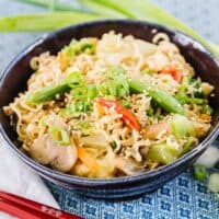 Ramen stir fry in dark bowl on blue tablecloth surrounded by red chopsticks and green onions