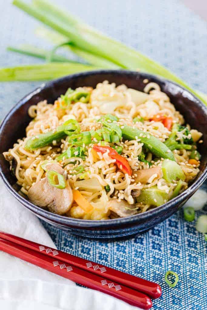 Ramen stir fry in dark bowl on blue tablecloth surrounded by red chopsticks and green onions