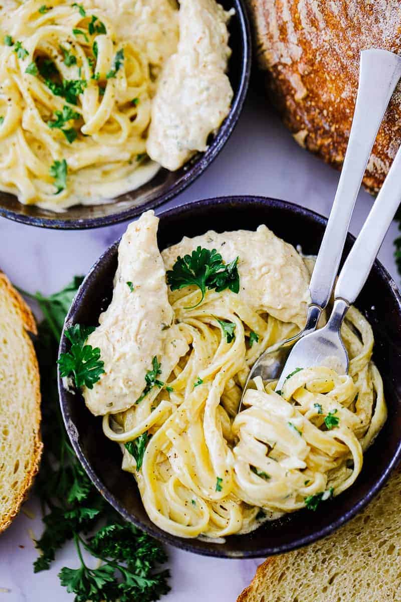 Top view of chicken alfredo in a dark bowl with fork and spoon surrounded by bread
