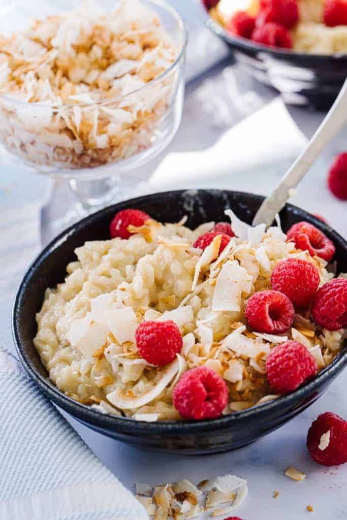 Rice pudding topped with coconut and raspberries in a dark bowl with a spoon dipped inside