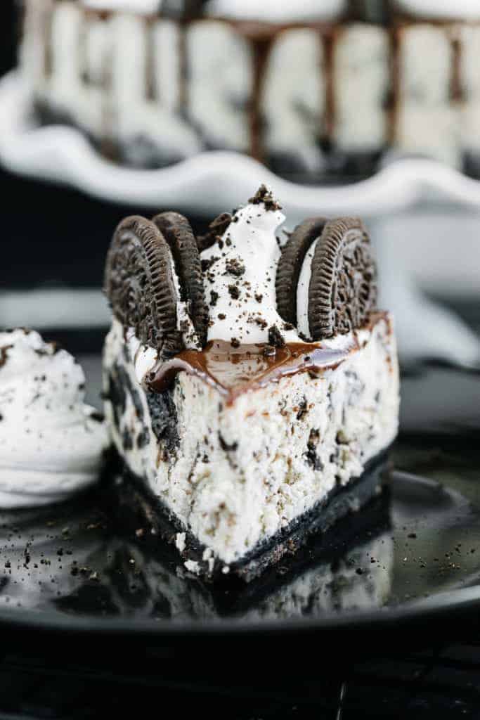 Slice of oreo cheesecake topped with chocolate drizzle, oreos, and cream on a dark plate