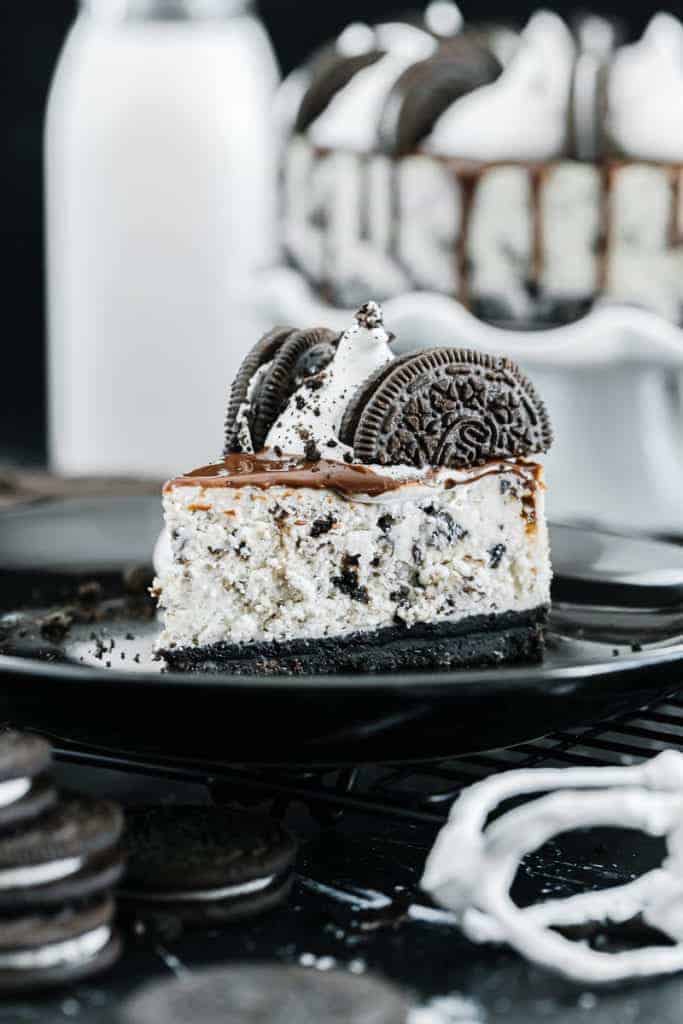 Slice of oreo cheesecake topped with chocolate drizzle, oreos, and cream on a dark plate surrounded by oreos and glass of milk