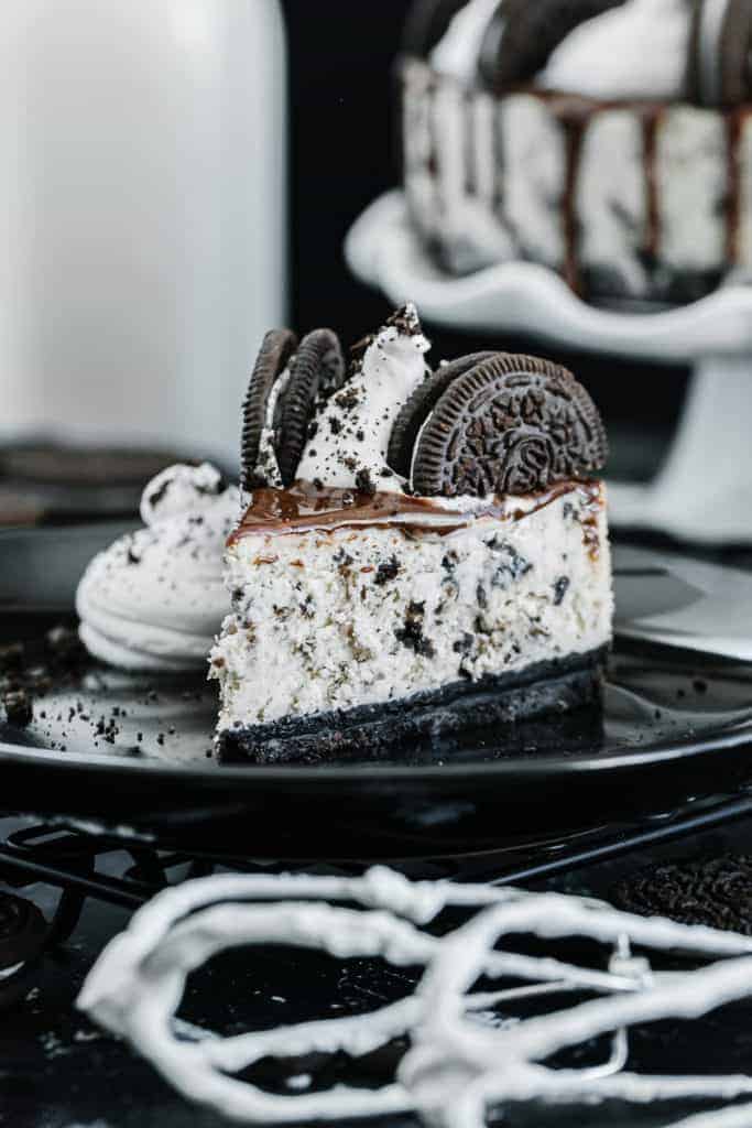 Slice of oreo cheesecake topped with chocolate drizzle, oreos, and cream on a dark plate surrounded by oreos and glass of milk