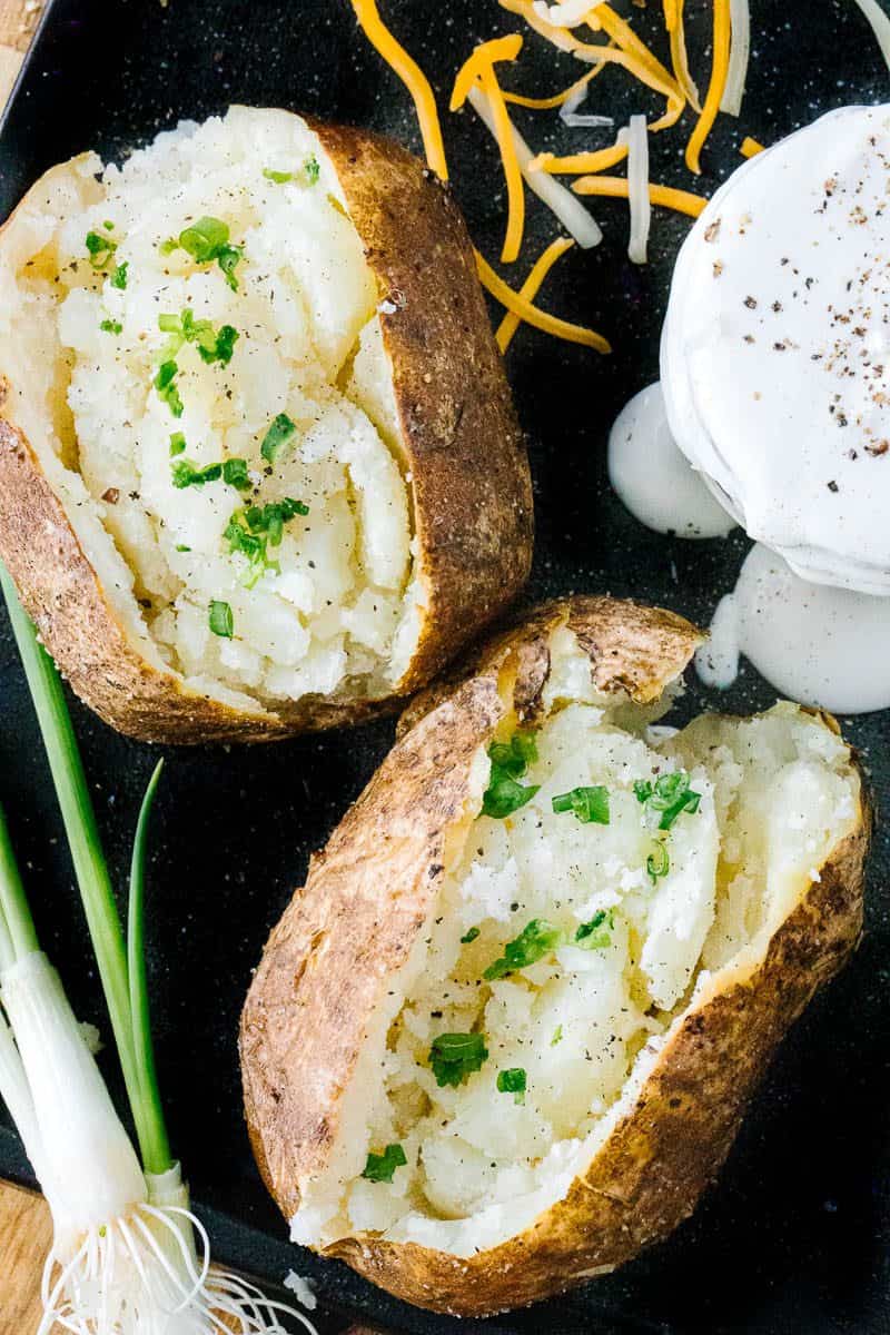 Top view of opened baked potatoes with scallions sprinkled on top. Next to sour cream and scallions