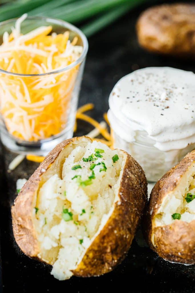 Baked potatoes sprinkled with scallions and next to a cup of shredded cheese and jar of sour cream