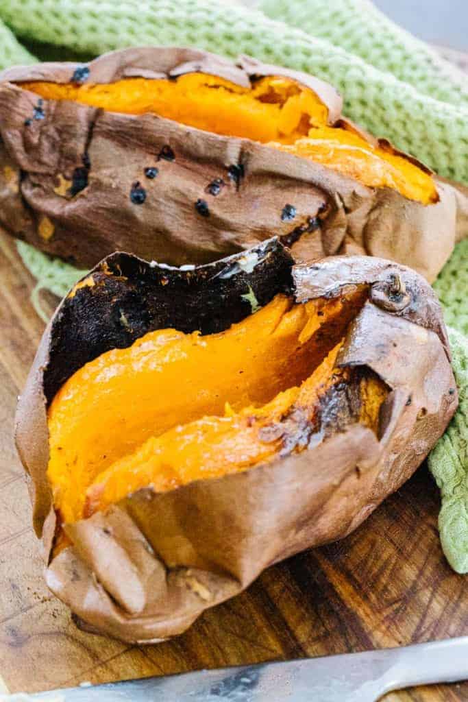 Two air fried sweet potatoes split open on a cutting board next to a green towel and buttered knife