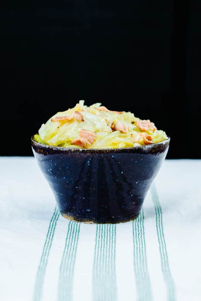 cooked cabbage and bacon pieces in dark bowl on white and green stripped towel