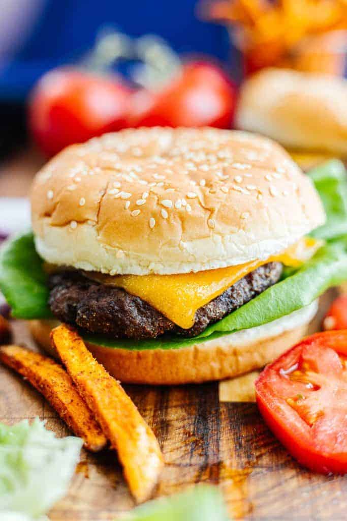 Side view close up of a frozen air fryer hamburger in bun with cheese and lettuce next to tomato slices and sweet potato fries