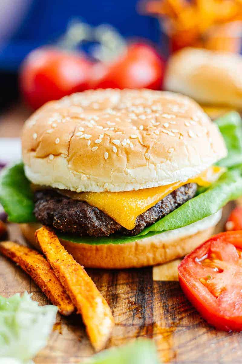 Side view close up of hamburger in bun with cheese and lettuce next to tomato slices and sweet potato fries