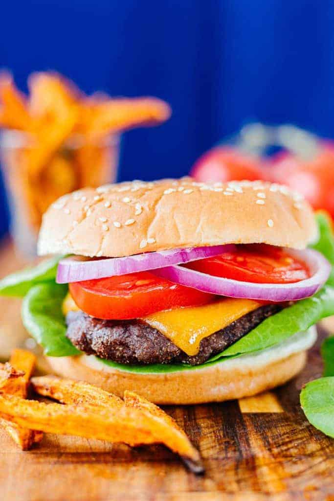 Close up of air fried hamburger in bun with cheese, tomato, onion, and lettuce next to sweet potato fries