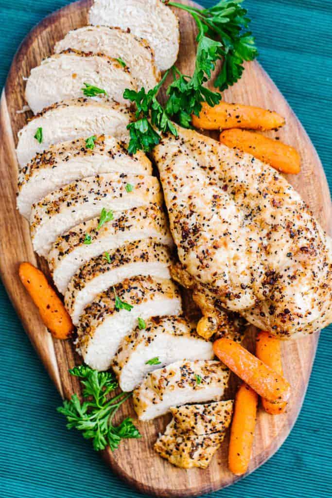 Top view of Air fryer chicken breast sliced and whole topped with seasoning and next to parsley and carrots all on a wooden board