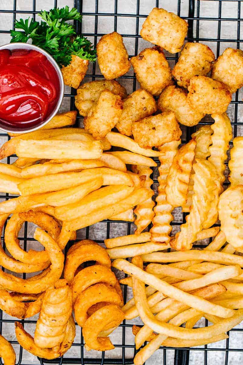 Top view of air fryer curly fries, crinkle fries, tater tots, and steak fries on black cooling rack next to cup of ketchup and parsley