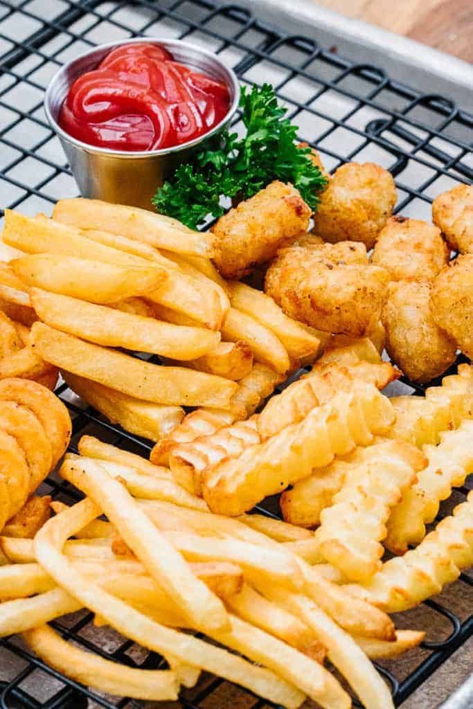 Side view of air fryer curly fries, crinkle fries, tater tots, and steak fries on black cooling rack next to cup of ketchup and parsley