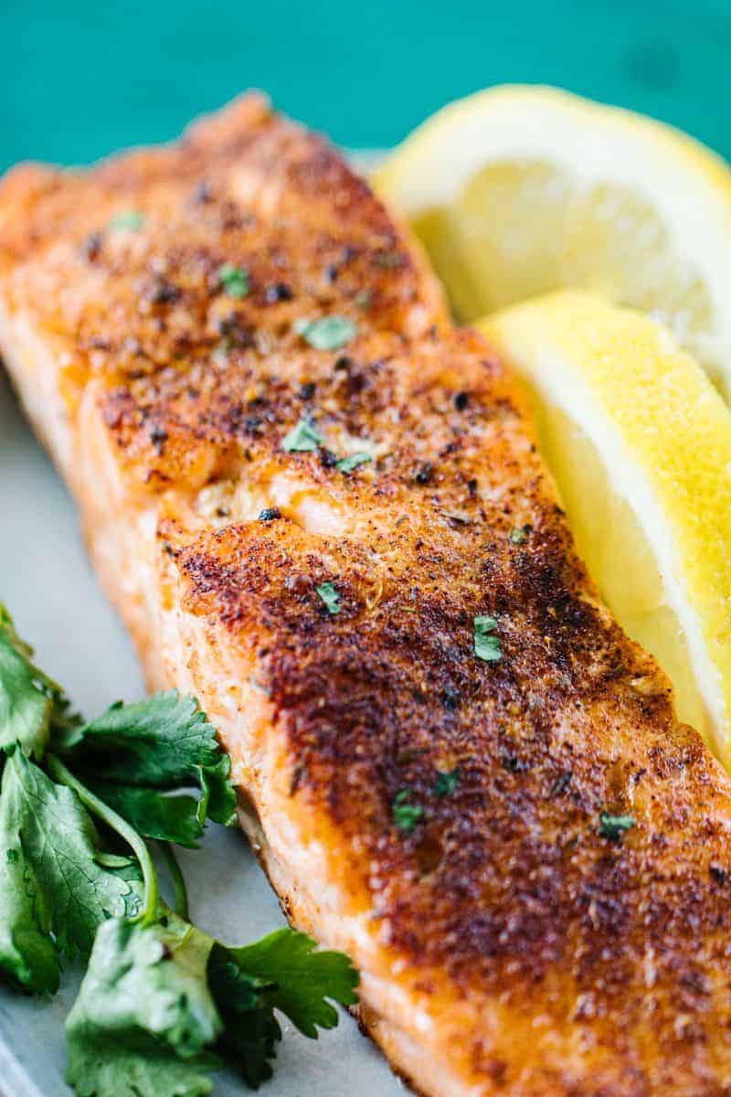 Close up of air fried salmon topped with parsley on a white plate next to lemon wedges and parsley next to a teal cloth