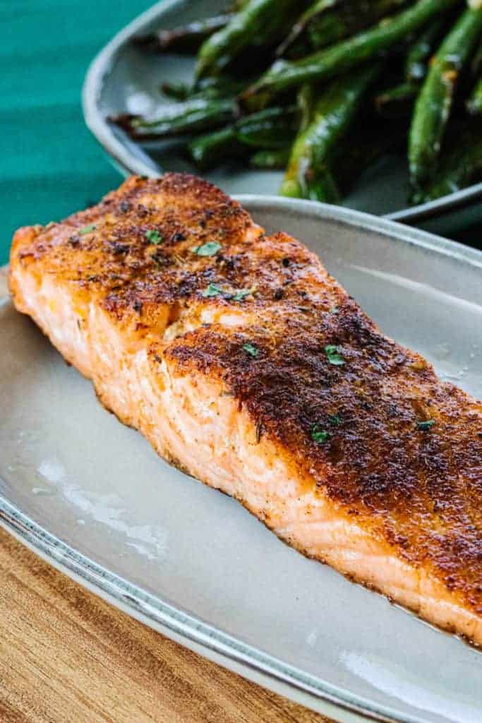 Air fried salmon topped with parsley on a white plate next to a teal cloth and a bowl of green beans