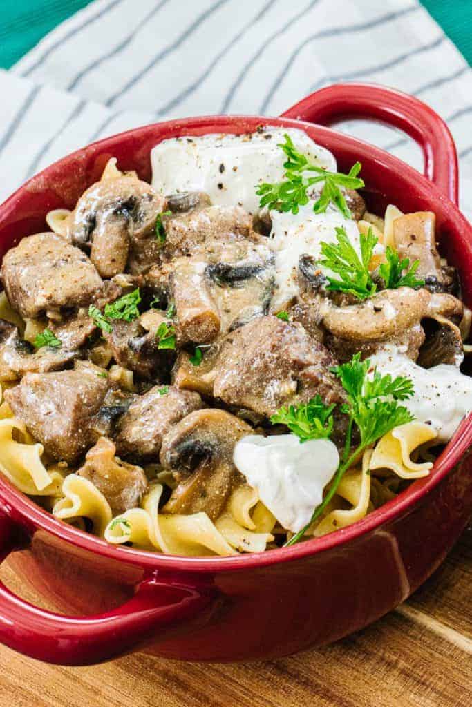Close up of instant pot beef stroganoff topped with parsley in red bowl next to white stripped cloth