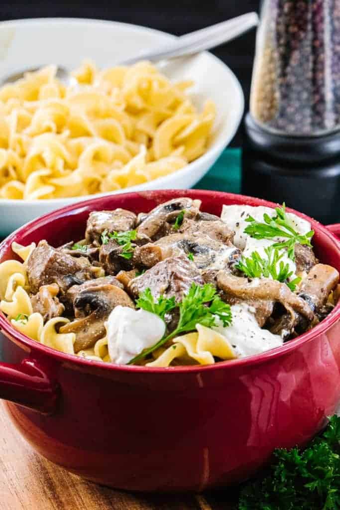 Instant pot beef stroganoff in red bowl in front of white bowl filled with cooked pasta next to peppercorn