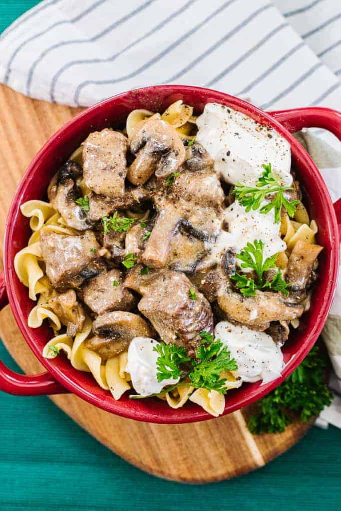 Top view of instant pot beef stroganoff topped with parsley and in a red bowl placed on a wooden board next to a white stripped cloth and teal cloth