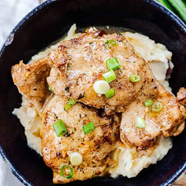 Close up of brown butter garlic chicken thighs in dark bowl on mashed potatoes and topped with green onions on top of woven mat next to white cloth and green onion stalk