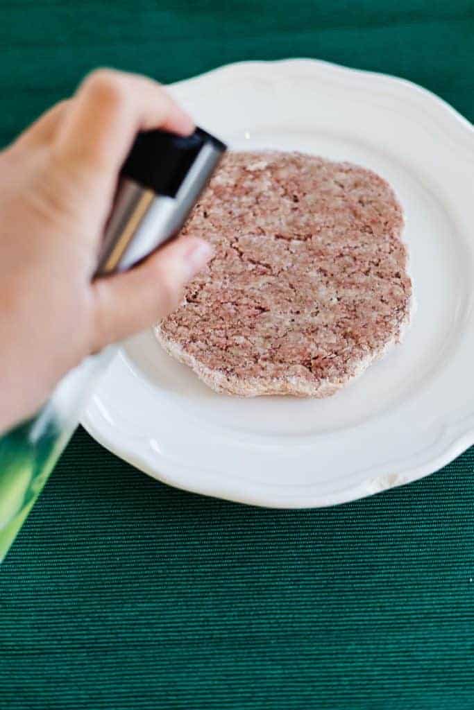 Raw frozen hamburger patty on white plate with hand misting patty with oil