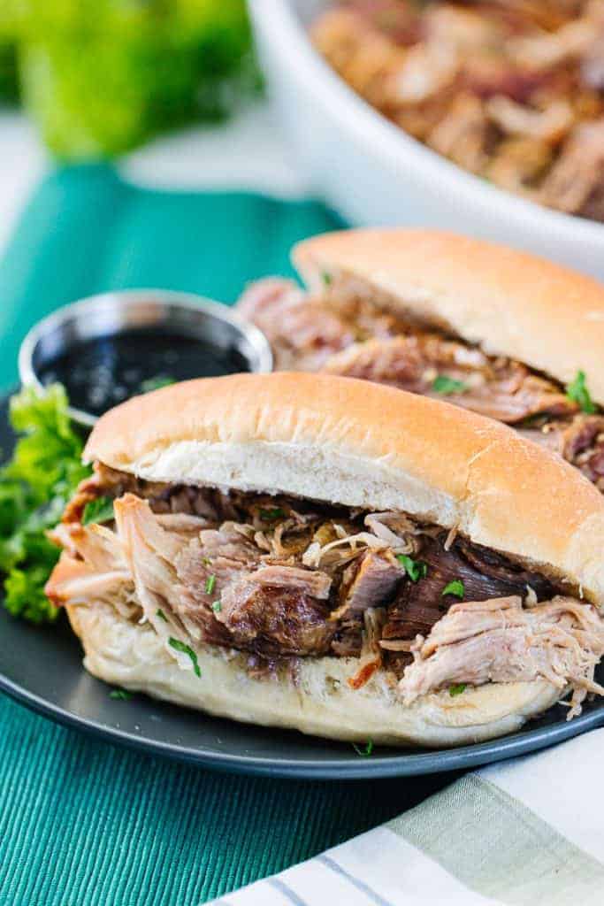 Instant pot pork roast in white bun on dark plate next to another sandwich and dipping sauce on a teal cloth next to bowl of shredded instant pot pork roast