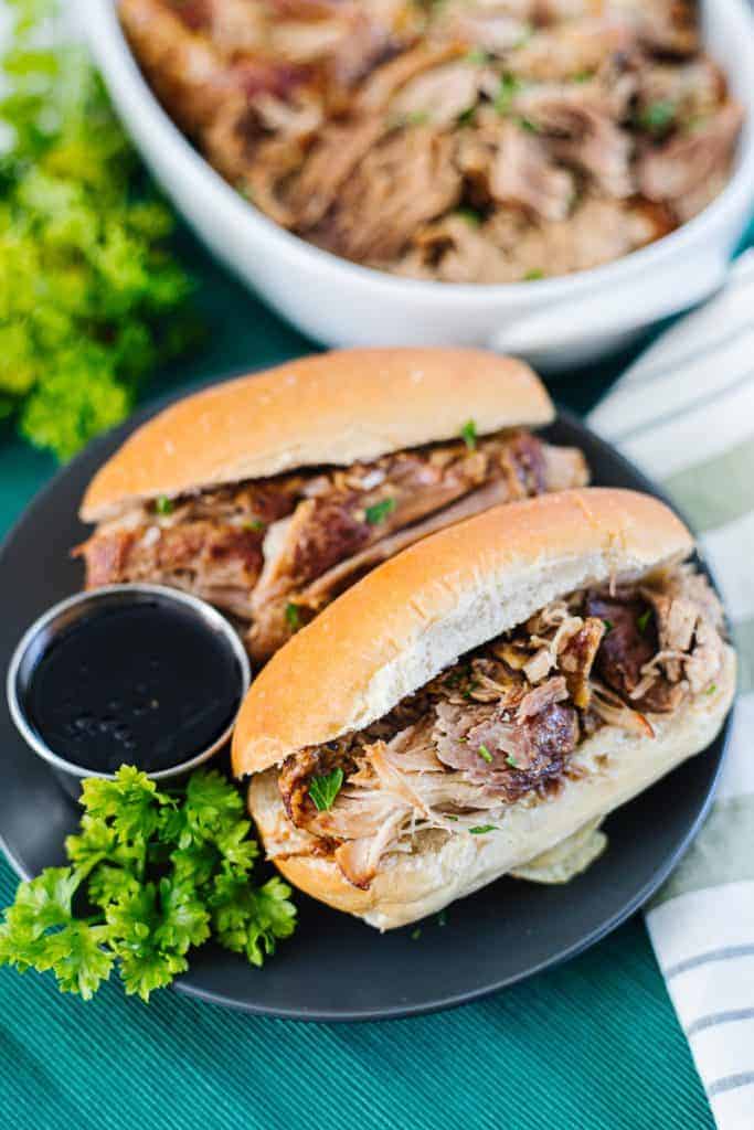 Top view of Instant pot pork roast in white bun on dark plate next to another sandwich and dipping sauce on a teal cloth next to bowl of shredded instant pot pork roast