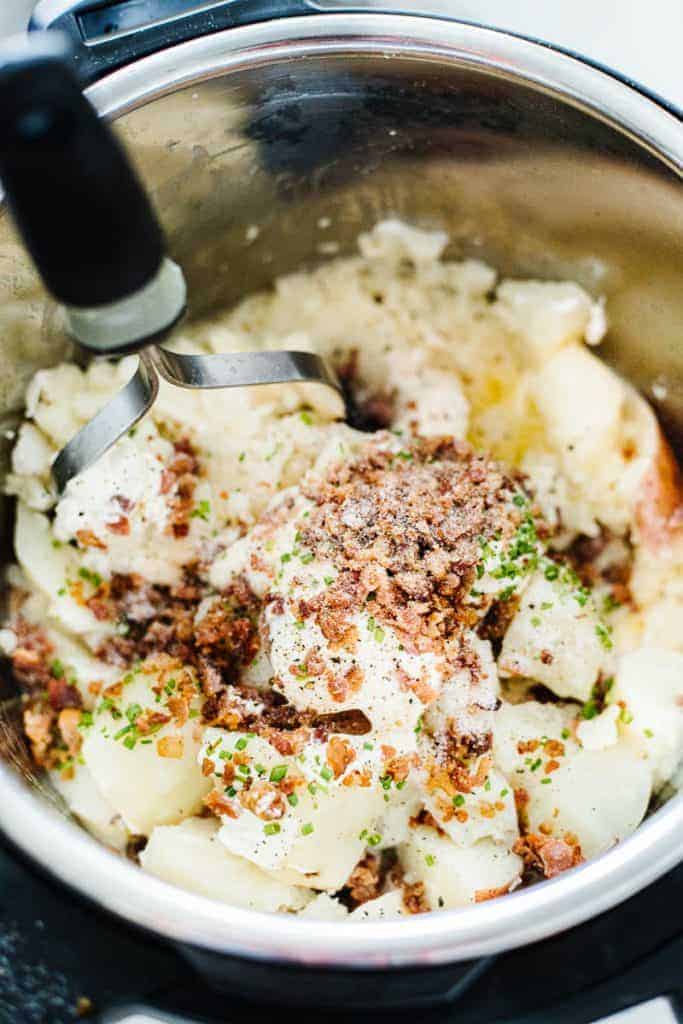 Top view of potatoes, bacon, sour cream, and green onions in instant pot being mashed with potato masher