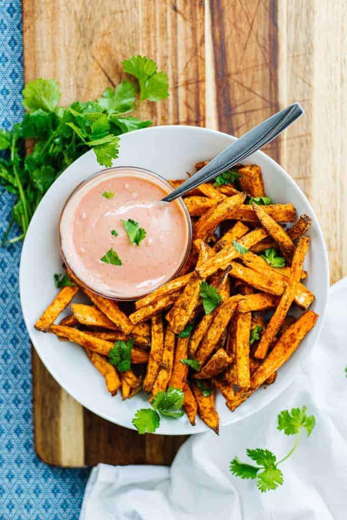 top view of sweet potato fries in white bowl topped with cilantro next to a bowl of dipping sauce on a cutting board, blue cloth and next to cilantro and a white napkin