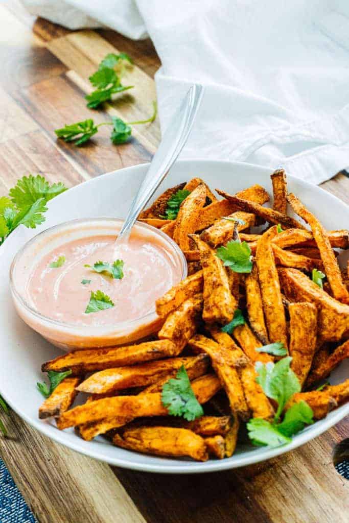 Close up of sweet potato fries white bowl topped with cilantro next to bowl of dipping sauce on top of a wood board