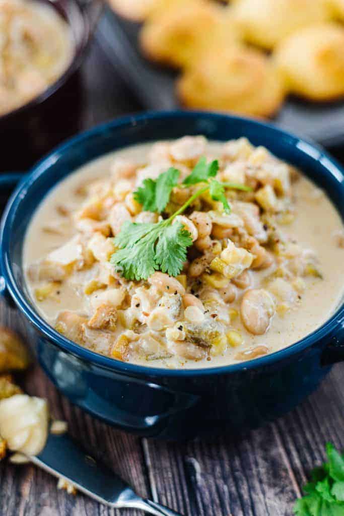 Close up of instant pot white chicken chili topped with cilantro and in a dark blue bowl in front of corn bread and next to a butter knife with butter