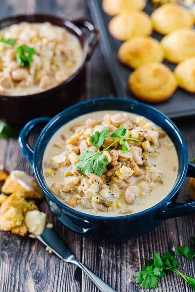 Instant pot white chicken chili topped with cilantro in dark blue bowl next to corn bread and buttered knife in front of another bowl of chili and muffin tin of cornbread