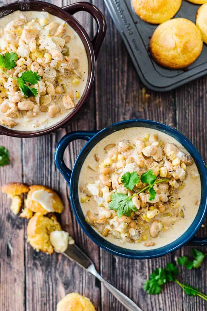 Top view of Instant pot white chicken chili topped with cilantro in dark blue bowl next to corn bread and buttered knife below of another bowl of chili and tin of cornbread