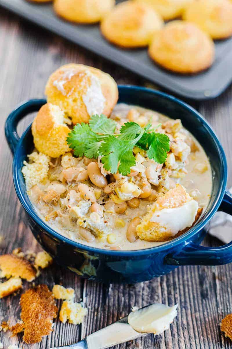 Instant pot white chicken chili topped with cilantro and buttered cornbread in a dark blue bowl next to a buttered knife and cornbread in front of a muffin tin of cornbread