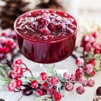 Instant pot cranberry sauce in glass container surrounded by frosted cranberries and pine cones on white cloth