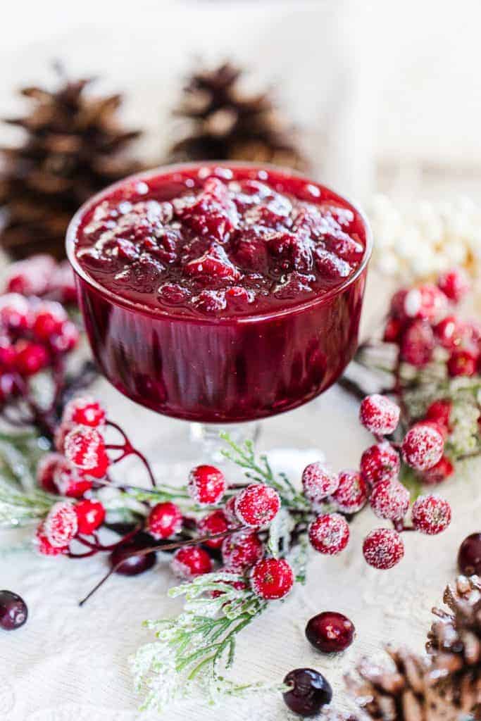 Instant pot cranberry sauce in glass container surrounded by frosted cranberries and pine cones on white cloth