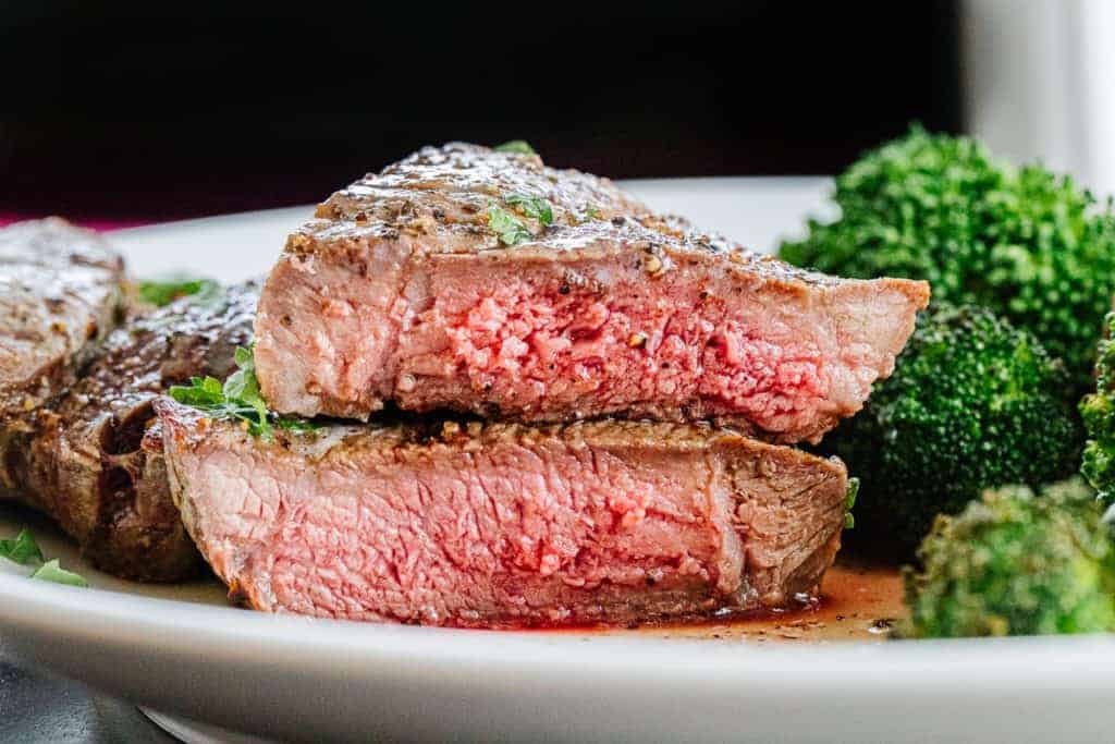 Close up side view of cut air fryer steak with one steak on top of another with pink centers and juice on white plate next to broccoli florets