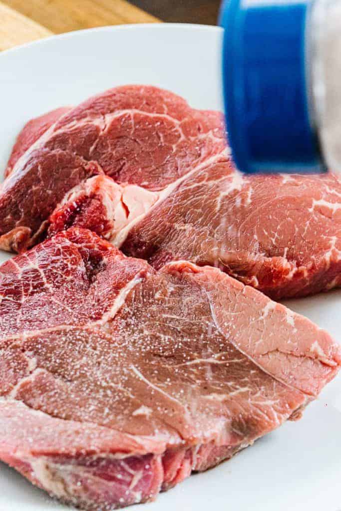 Raw steak pieces on a white plate with seasoning being sprinkled on top