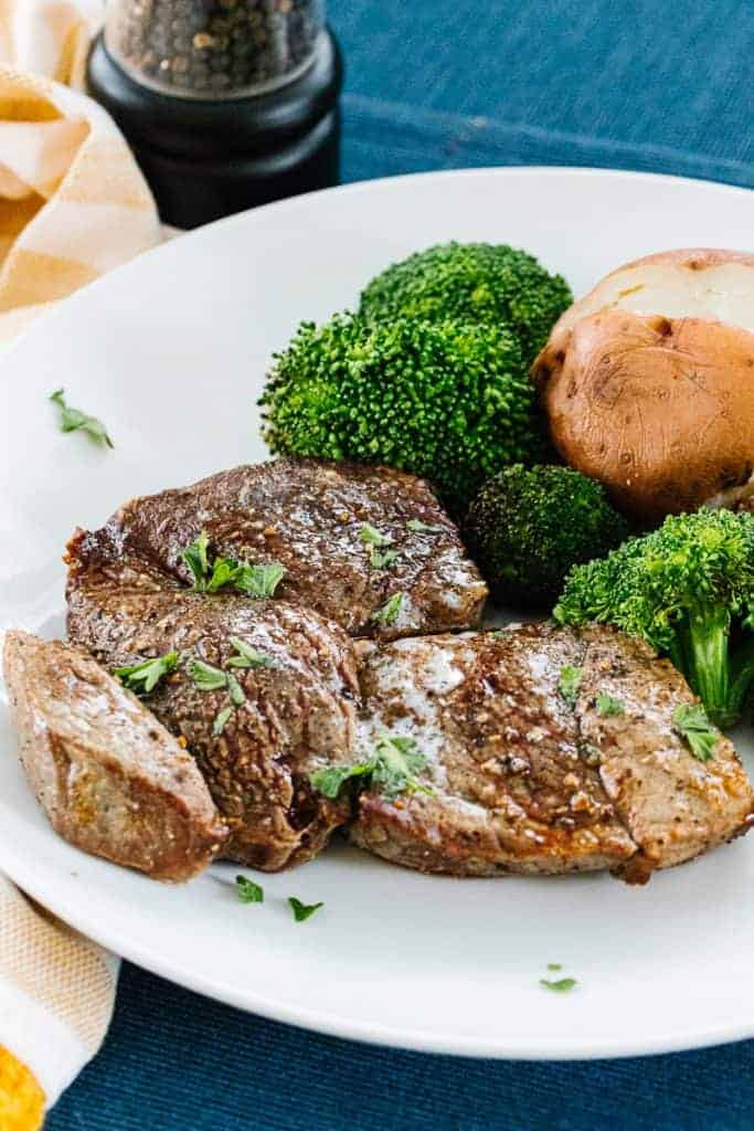 Air Fryer Steak on white plate topped with butter and parsley next to broccoli florets and a potato all on a blue cloth and yellow striped cloth