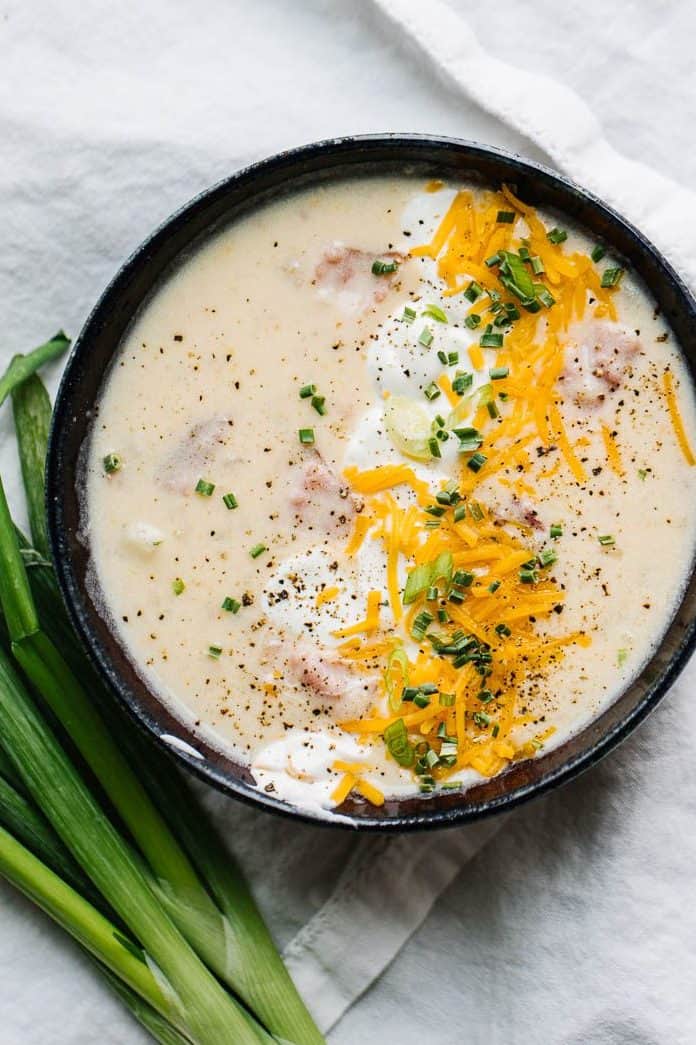 Top view of potato ham soup topped with cheese, chives, and sour cream on white cloth next to green onions