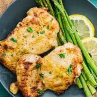 top view of air fryer chicken thighs and cooked asparagus next to lemons on a plate on an oblong wood plate and teal cloth