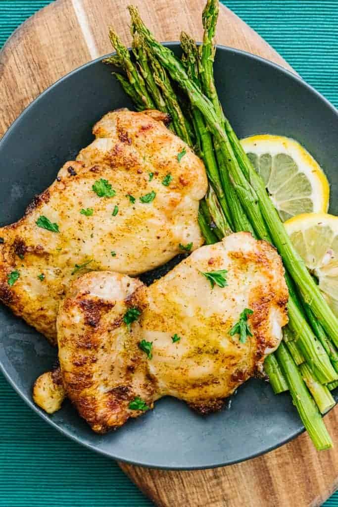 top view of air fryer chicken thighs and cooked asparagus next to lemons on a plate on an oblong wood plate and teal cloth