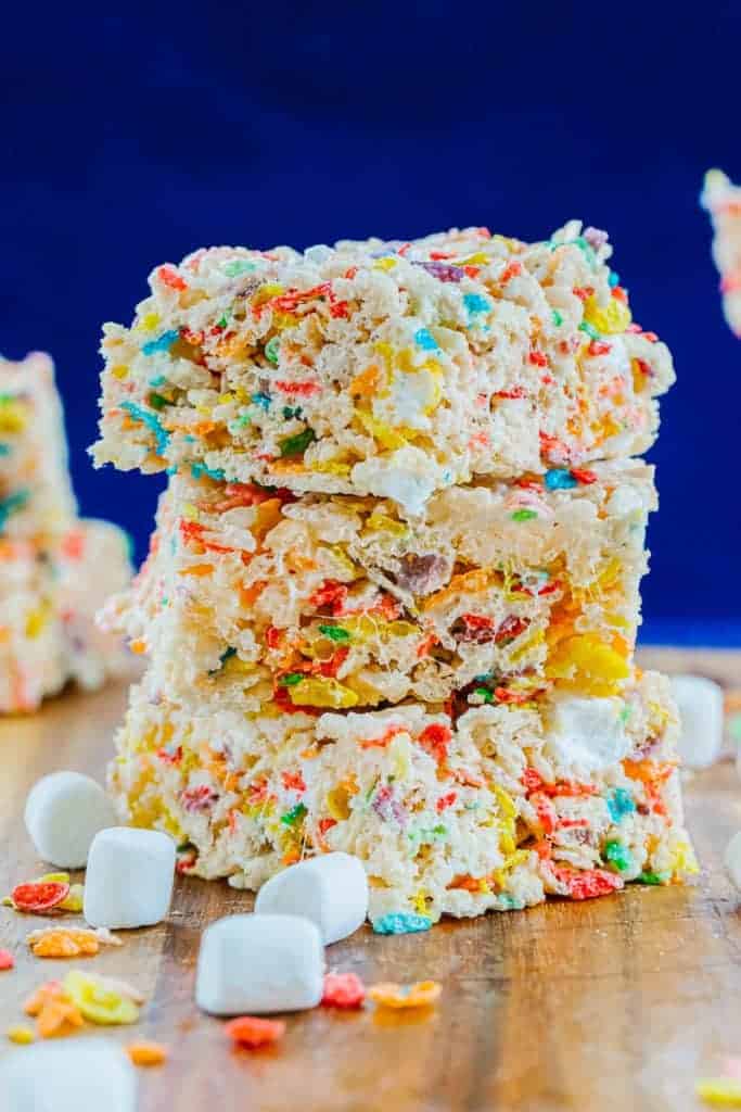 three fruity pebble rice krispies stacked on top of each other on a wood board surrounded by marshmallows and fruity pebbles with a blue background