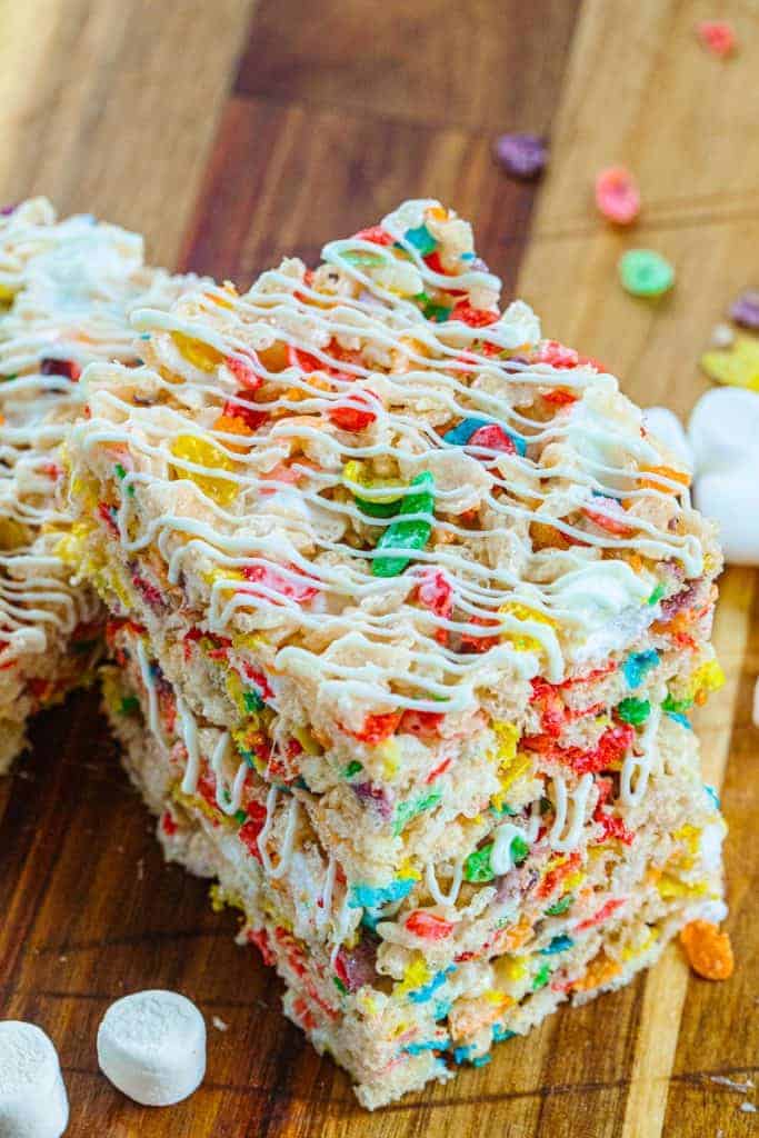 fruity pebble rice krispies squares stacked drizzled with white icing on a wood board surrounded by marshmallows and fruity pebbles