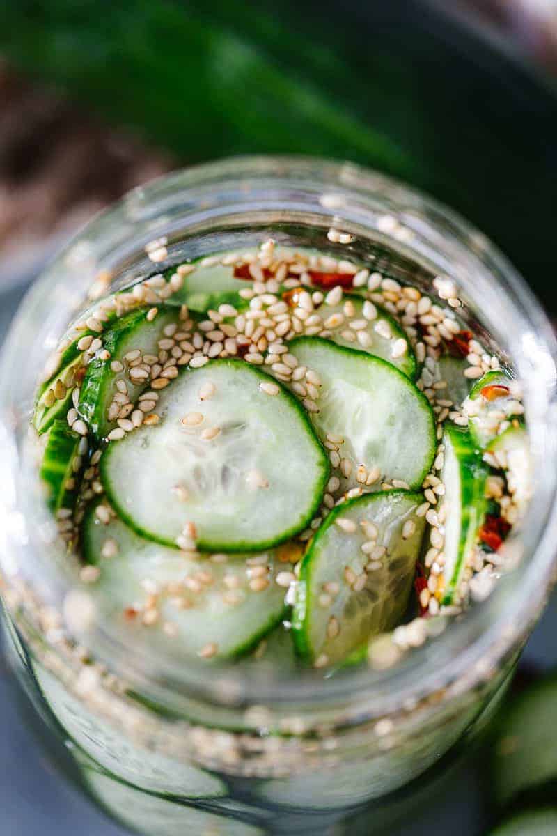 Top view of sliced cucumbers in liquid with sesame seeds in glass jar